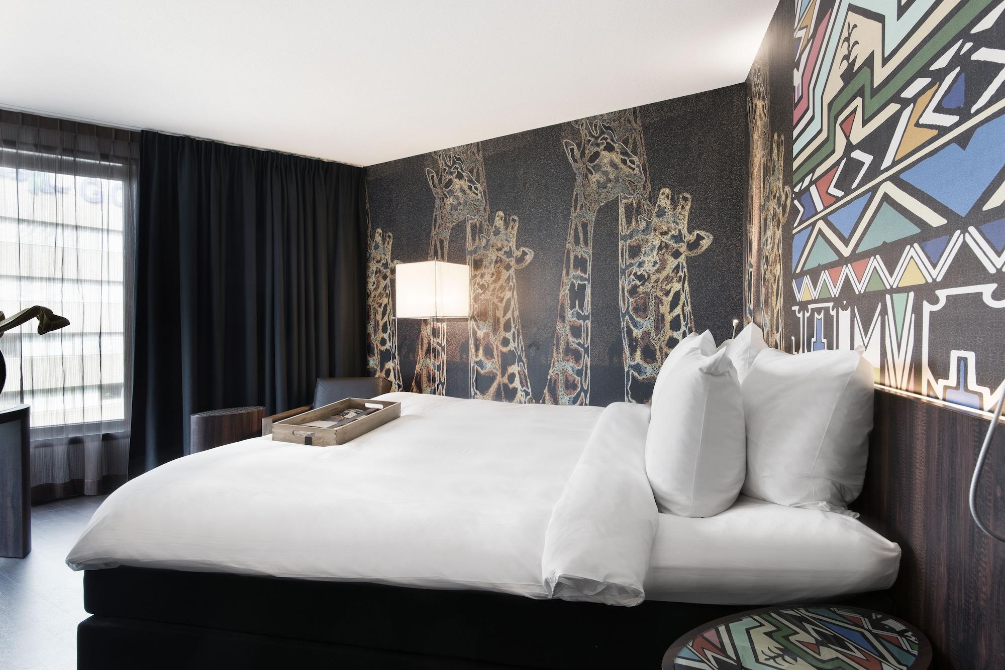 Mainport Hotel Rotterdam, A Hilton Affiliate Hotel Экстерьер фото The photo shows a modern hotel room featuring a large bed with white linens and several pillows. The backdrop includes a striking wall with artistic representations of giraffes, adding a unique, thematic touch. Next to the bed, there is a wooden tray