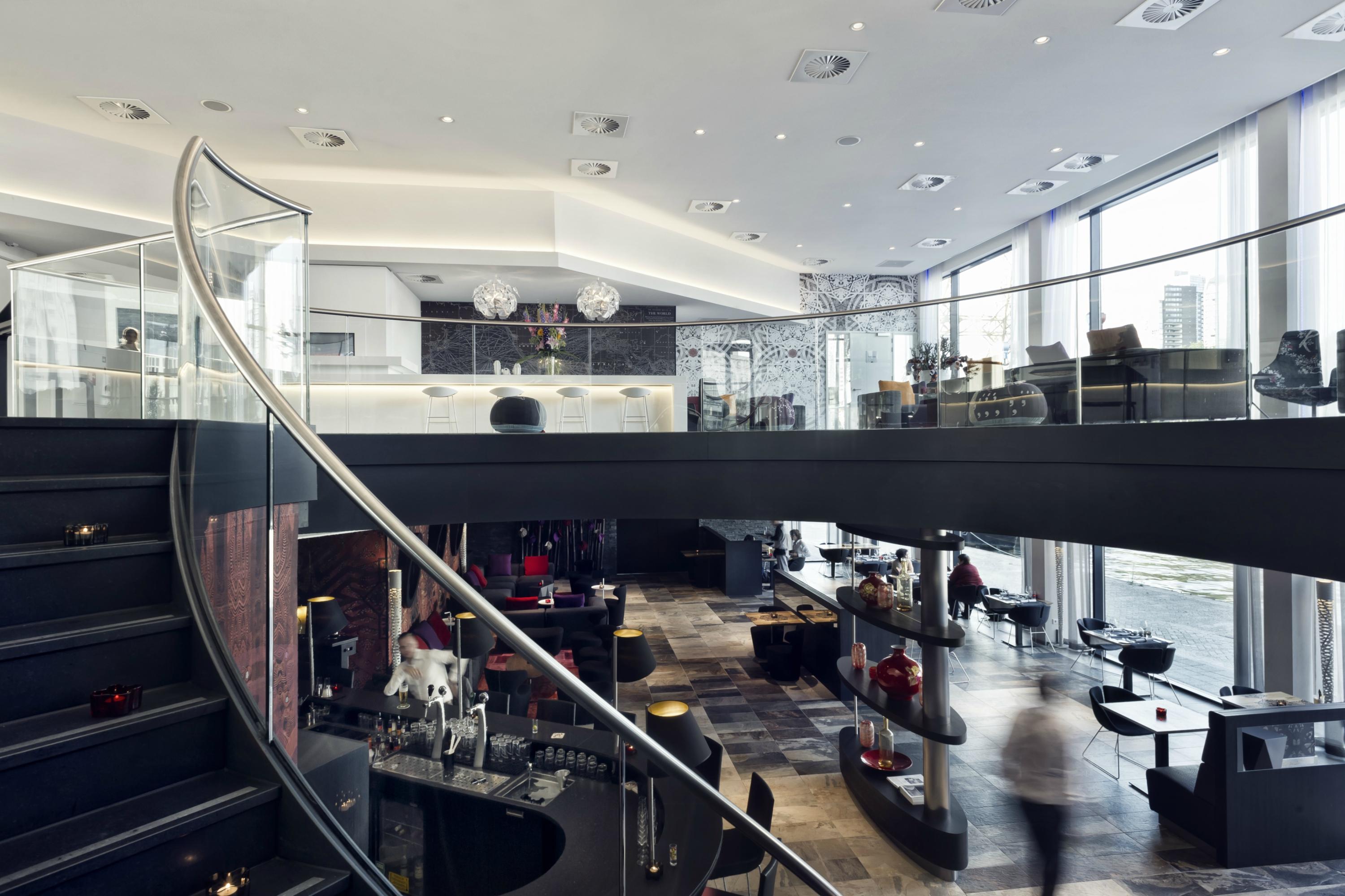 Mainport Hotel Rotterdam, A Hilton Affiliate Hotel Экстерьер фото The photo shows a modern restaurant or lounge area with a sleek and sophisticated design. It features a large, open space with high ceilings and large windows that allow natural light to flood in. A curved staircase leads to a second level, creating 
