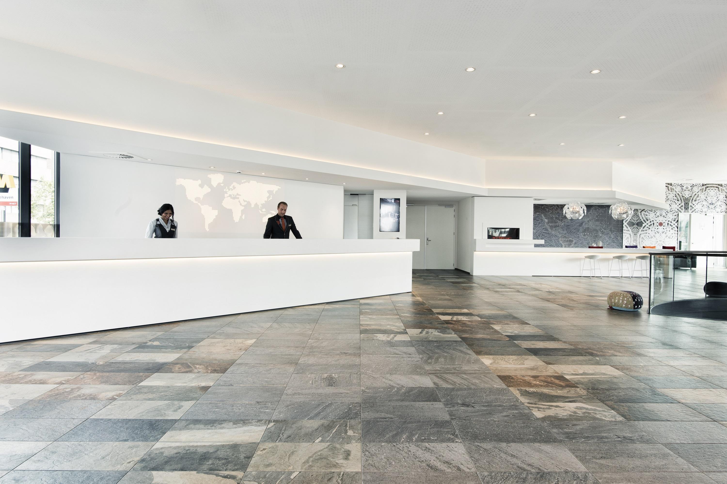 Mainport Hotel Rotterdam, A Hilton Affiliate Hotel Экстерьер фото The photo shows a modern reception area with a sleek and minimalist design. There is a long, white reception desk at the center, behind which two people are standing. The walls are mostly white, with subtle lighting integrated into the design, contri