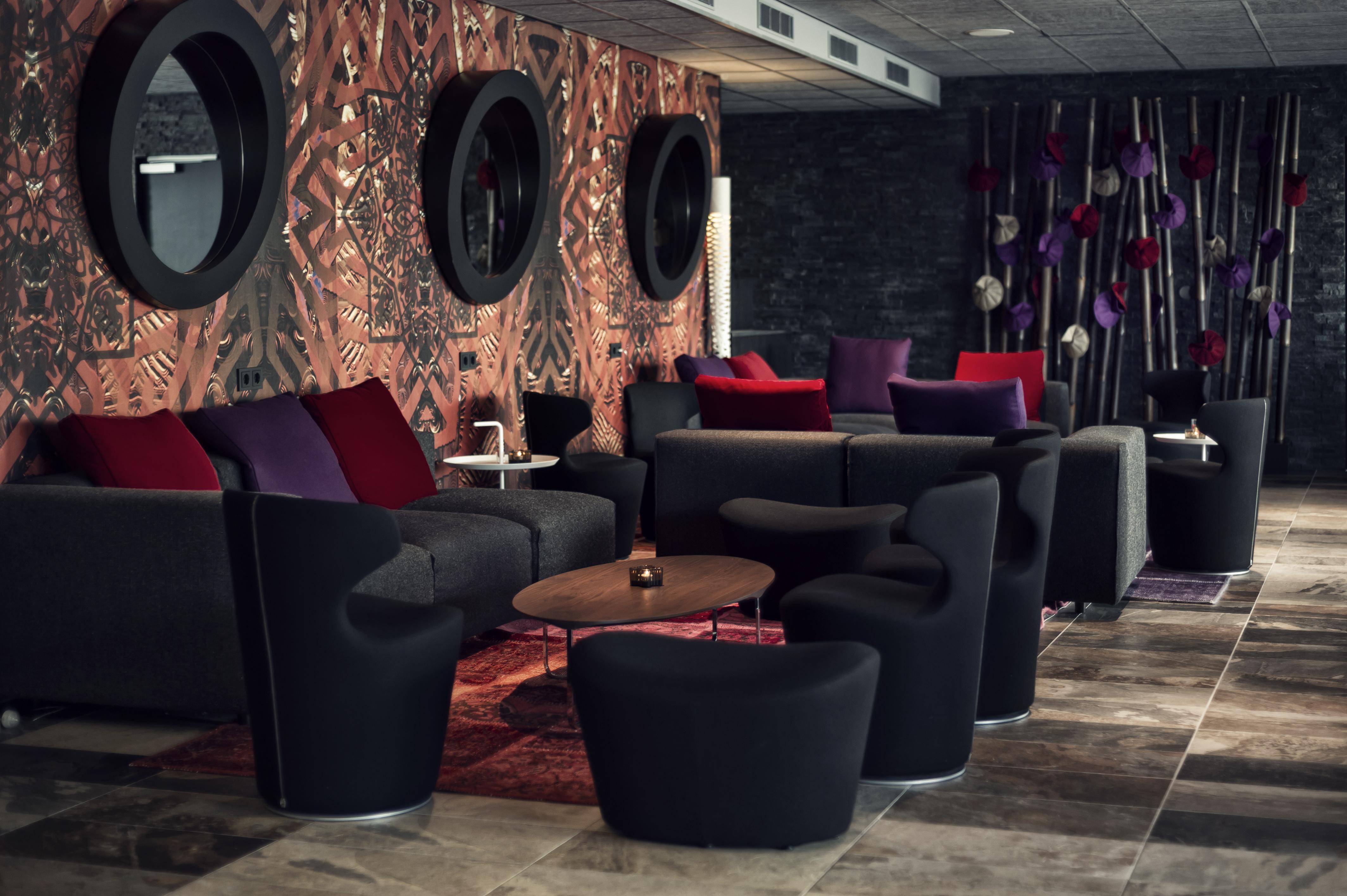 Mainport Hotel Rotterdam, A Hilton Affiliate Hotel Экстерьер фото The photo shows a modern lounge area featuring various seating arrangements. There are several dark, contemporary chairs and sofas, some adorned with colorful cushions in shades of purple and red. The background has a patterned wall with round mirror
