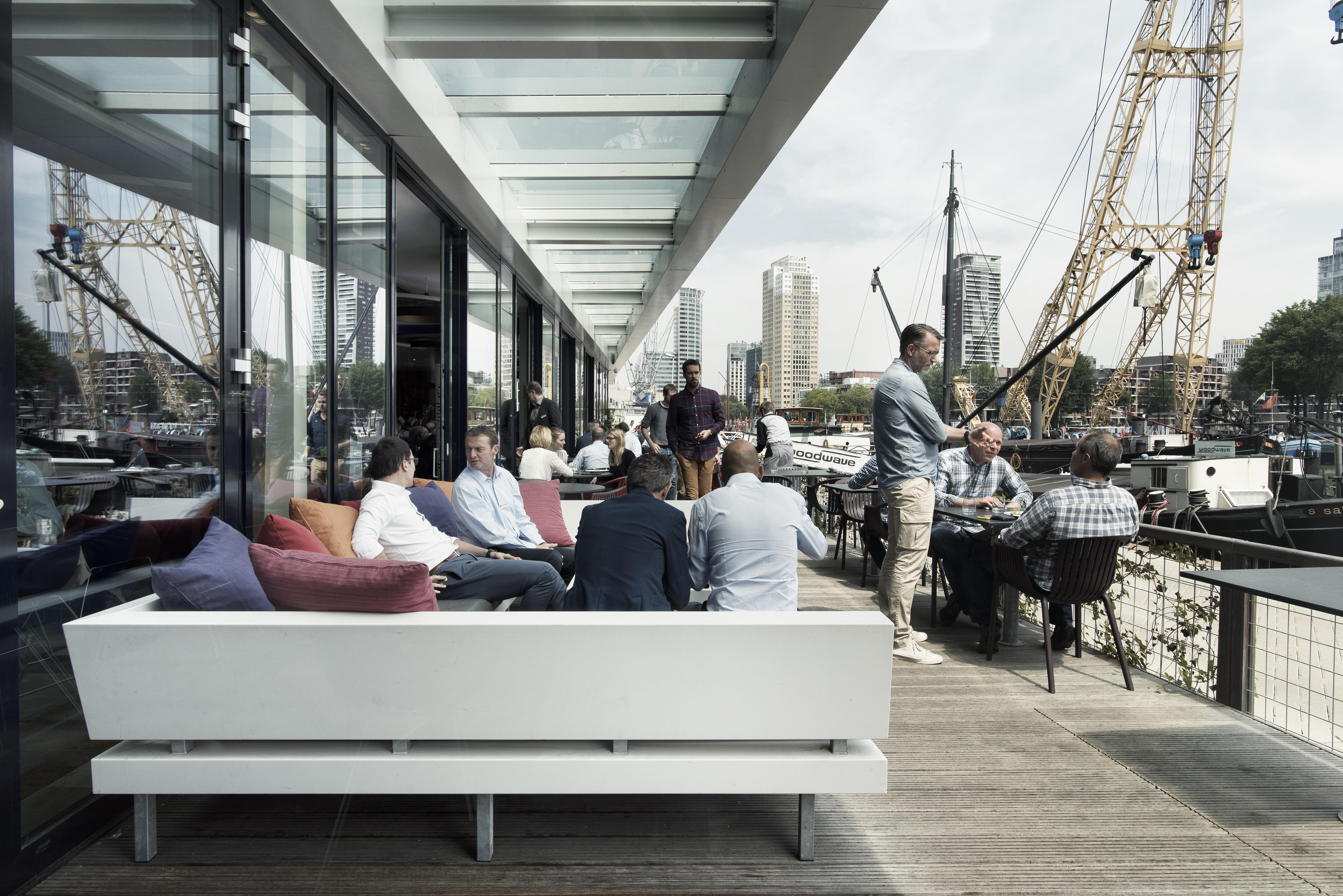 Mainport Hotel Rotterdam, A Hilton Affiliate Hotel Экстерьер фото The photo depicts an outdoor dining or gathering area that features a modern and stylish design. In the foreground, there is a white bench with colorful cushions, where several individuals are seated. They are dressed in business attire, suggesting a