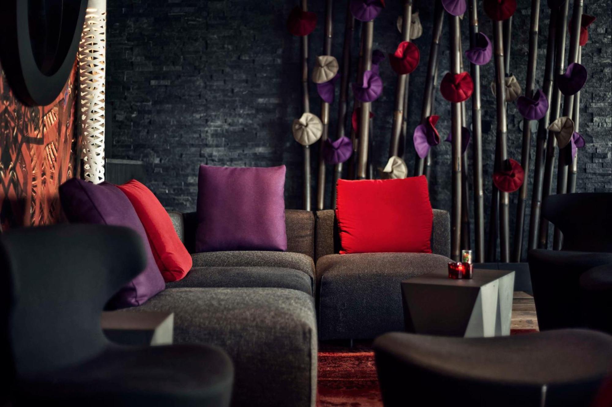 Mainport Hotel Rotterdam, A Hilton Affiliate Hotel Экстерьер фото The photo shows a stylish lounge area with modern seating. There is a dark-themed backdrop featuring textured black walls. A low, dark couch with multiple sections is adorned with colorful cushions, including deep purple and bright red pillows that a